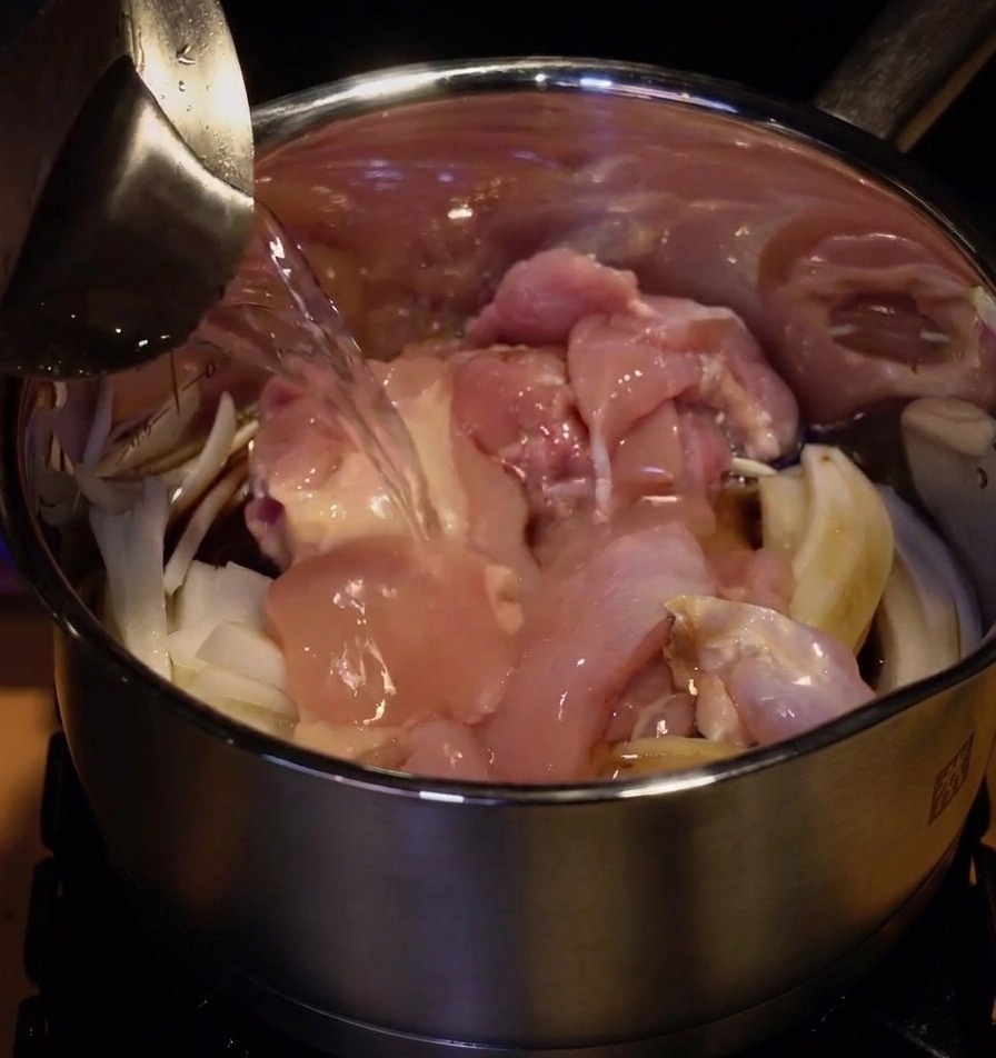 Acqua versata da una caraffa di metallo in un pentolino dacciaio pieno di pollo crudo e cipolle affettate.