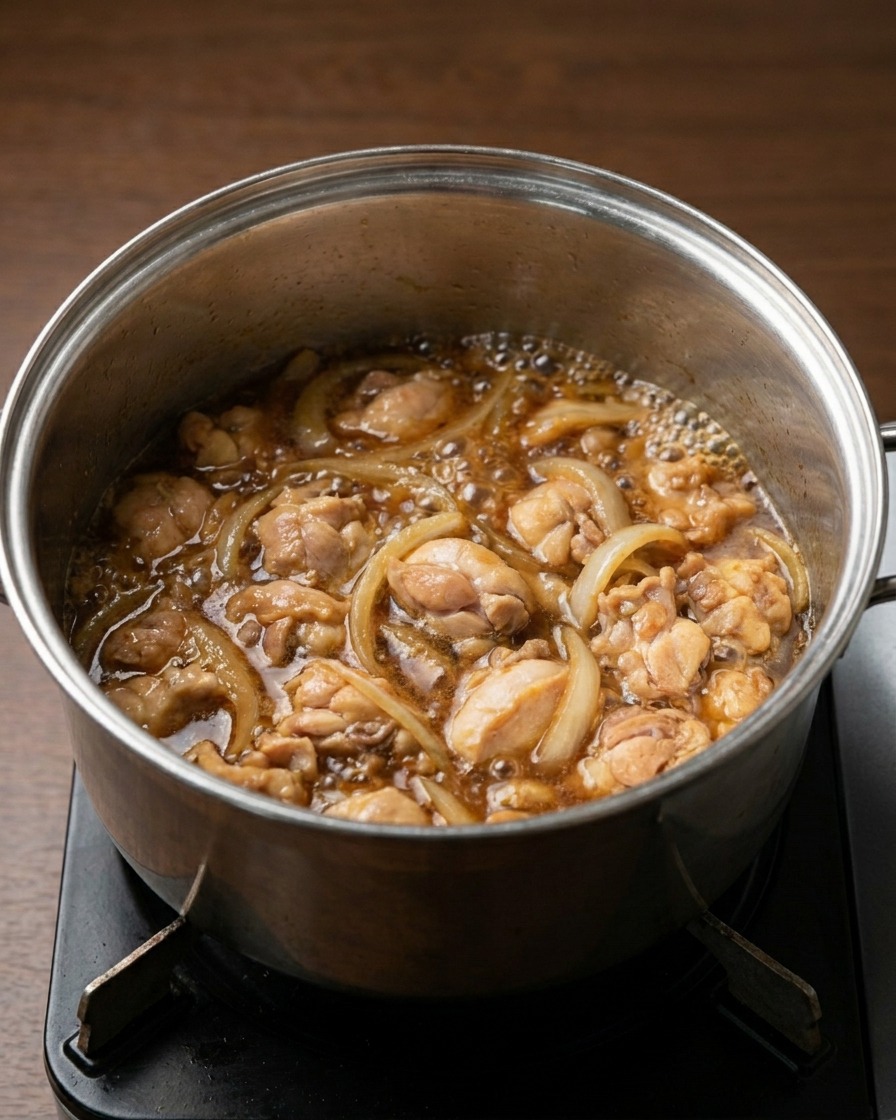 Un pentolino sul fuoco con pezzi di pollo e fette di cipolla che sobbollono in un liquido saporito marrone scuro.