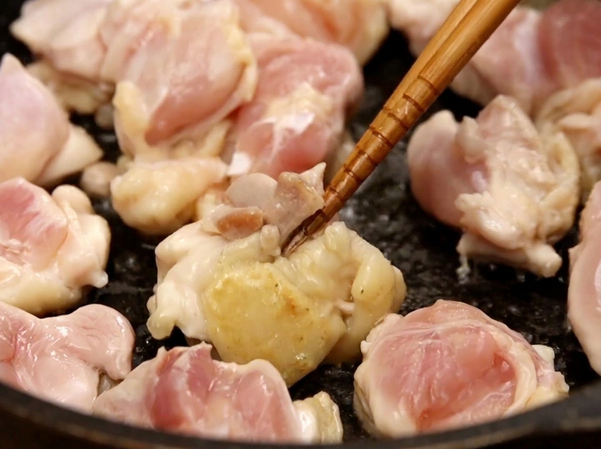 Pezzi di sovracoscia di pollo rosolati in una padella scura con bacchette di legno, con alcuni pezzi che iniziano a dorarsi.