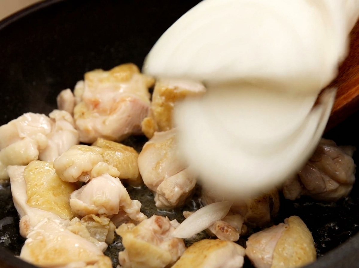 Aggiunta di fette di cipolla bianca in una padella con pezzi di pollo rosolati, mescolando il tutto.