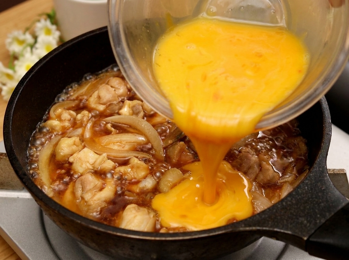 Un rivolo di uovo sbattuto versato da una ciotola di vetro in una padella con pollo e cipolle che sobbollono in una salsa scura.