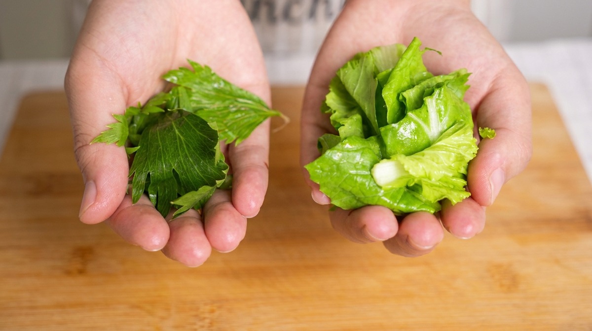 Due mani che tengono manciate separate di foglie di sedano tritate e baby bok choy sopra un tagliere di bambù.