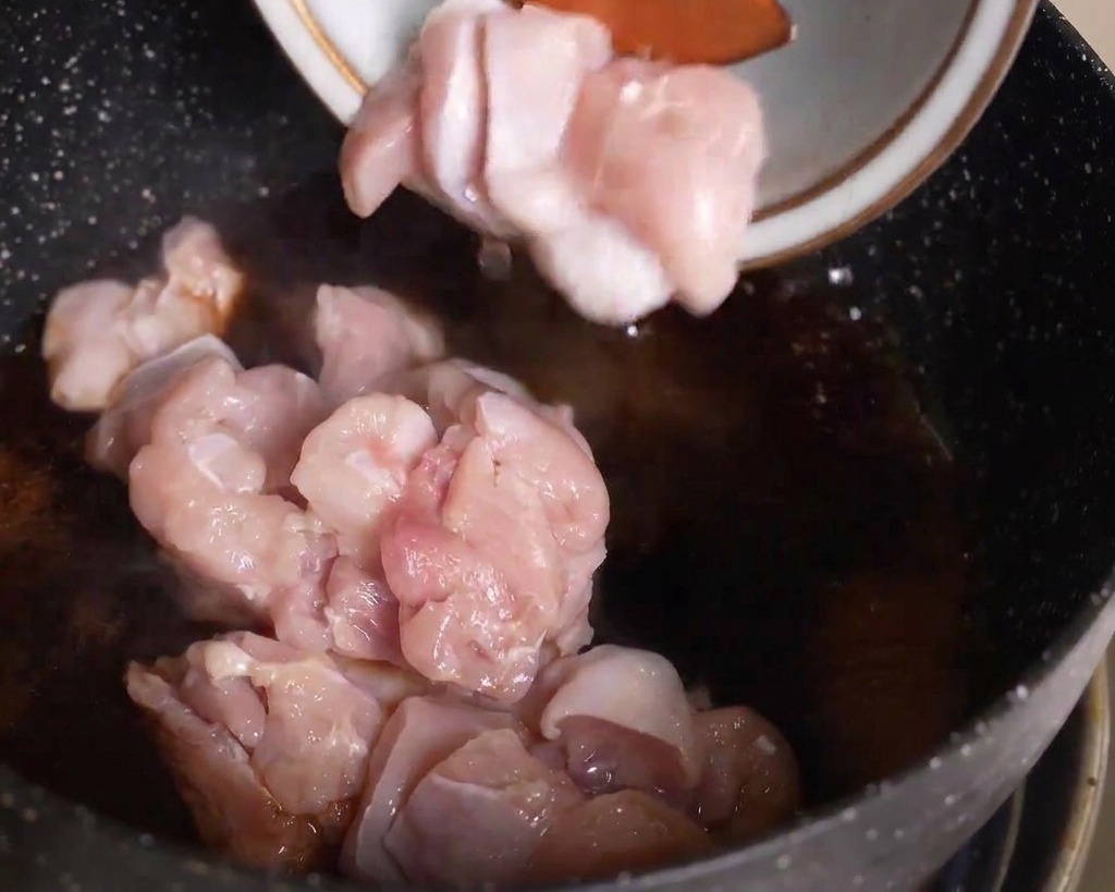 Pezzi di pollo crudo affettati aggiunti da una ciotola in una padella con salsa scura che sobbolle.