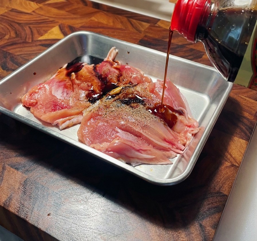 Salsa di soia versata da una bottiglia su sovracosce di pollo pepate in un vassoio dacciaio.