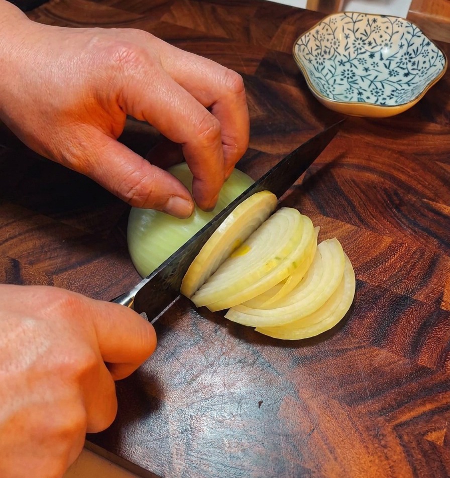 Mani di uno chef che usano un coltello per affettare una cipolla dorata in strisce sottili su un tagliere di legno scuro.