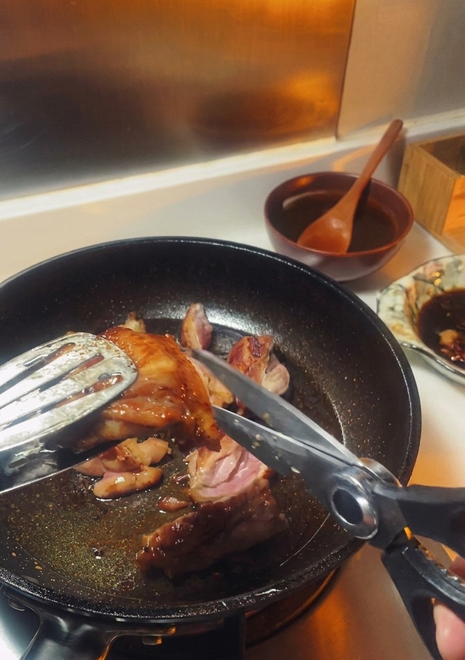 Forbici da cucina che tagliano una sovracoscia di pollo rosolata tenuta con pinze di metallo in padella.