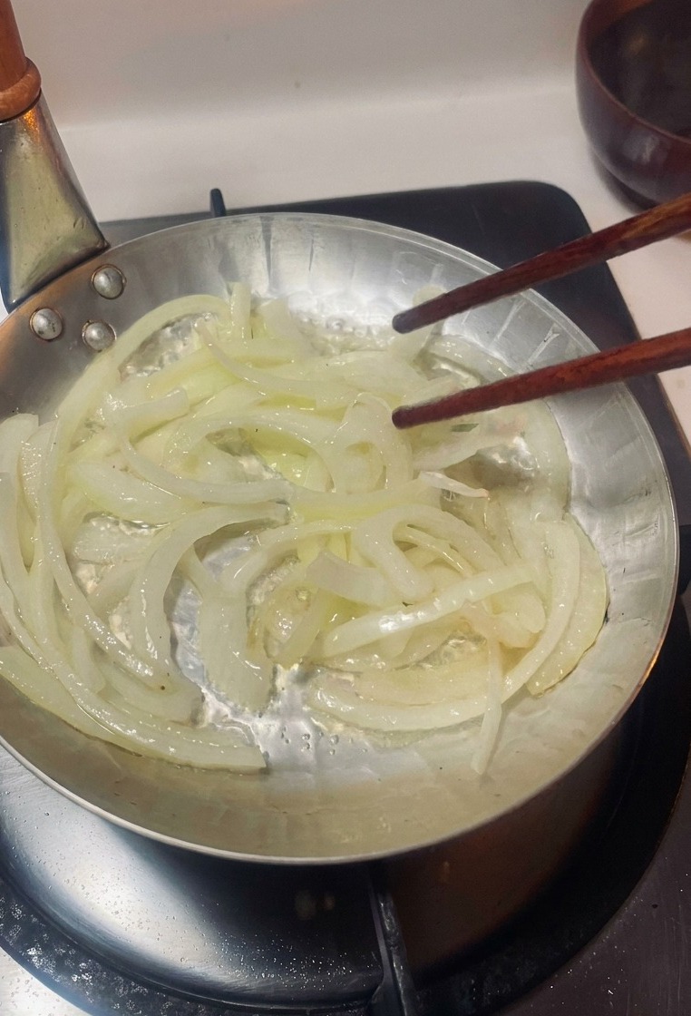 Cipolle affettate sottilmente mescolate con bacchette di legno in una piccola padella dargento su un fornello.