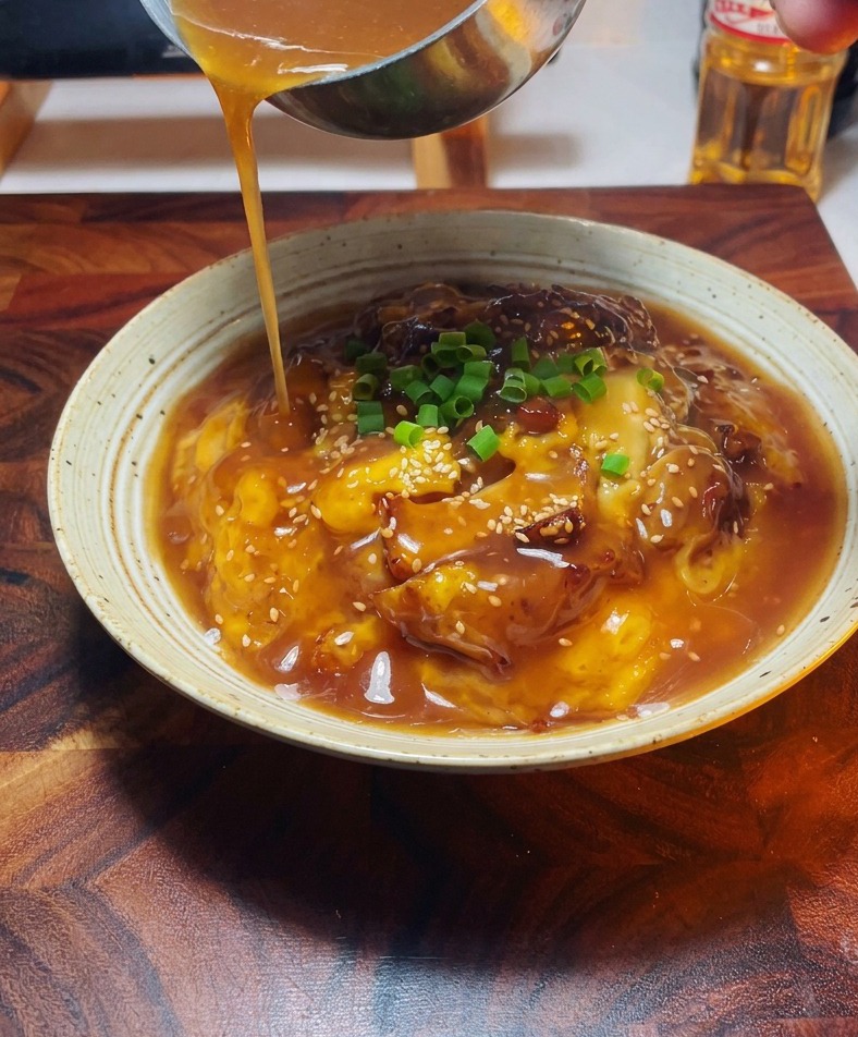 Dettaglio di una mano che usa un mestolo di metallo per versare una salsa dorata su una ciotola di pollo e uova guarnita.