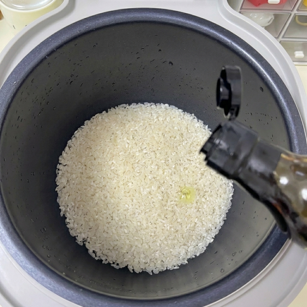 Una mano versa un filo di olio trasparente da una bottiglia scura in una pentola per cuociriso piena di riso bianco.