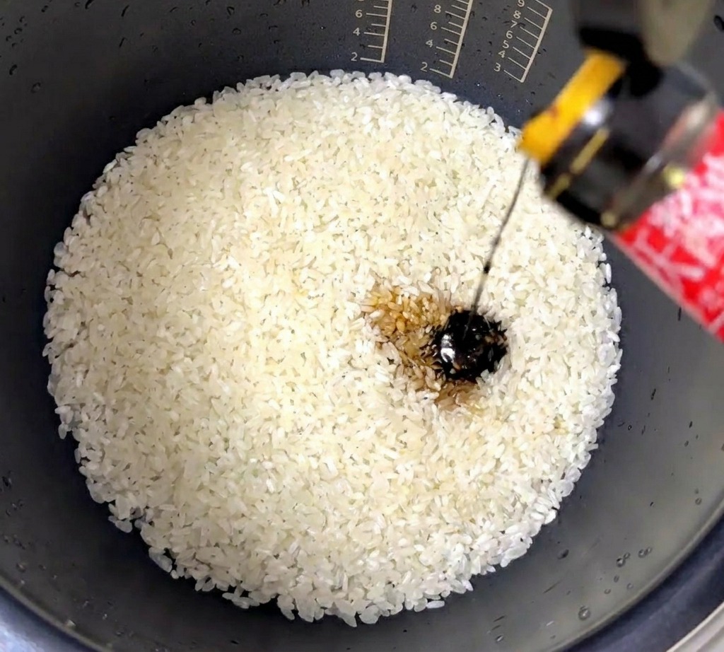 Un filo di salsa dostriche densa e scura viene versato in una pentola per cuociriso piena di riso bianco lavato.