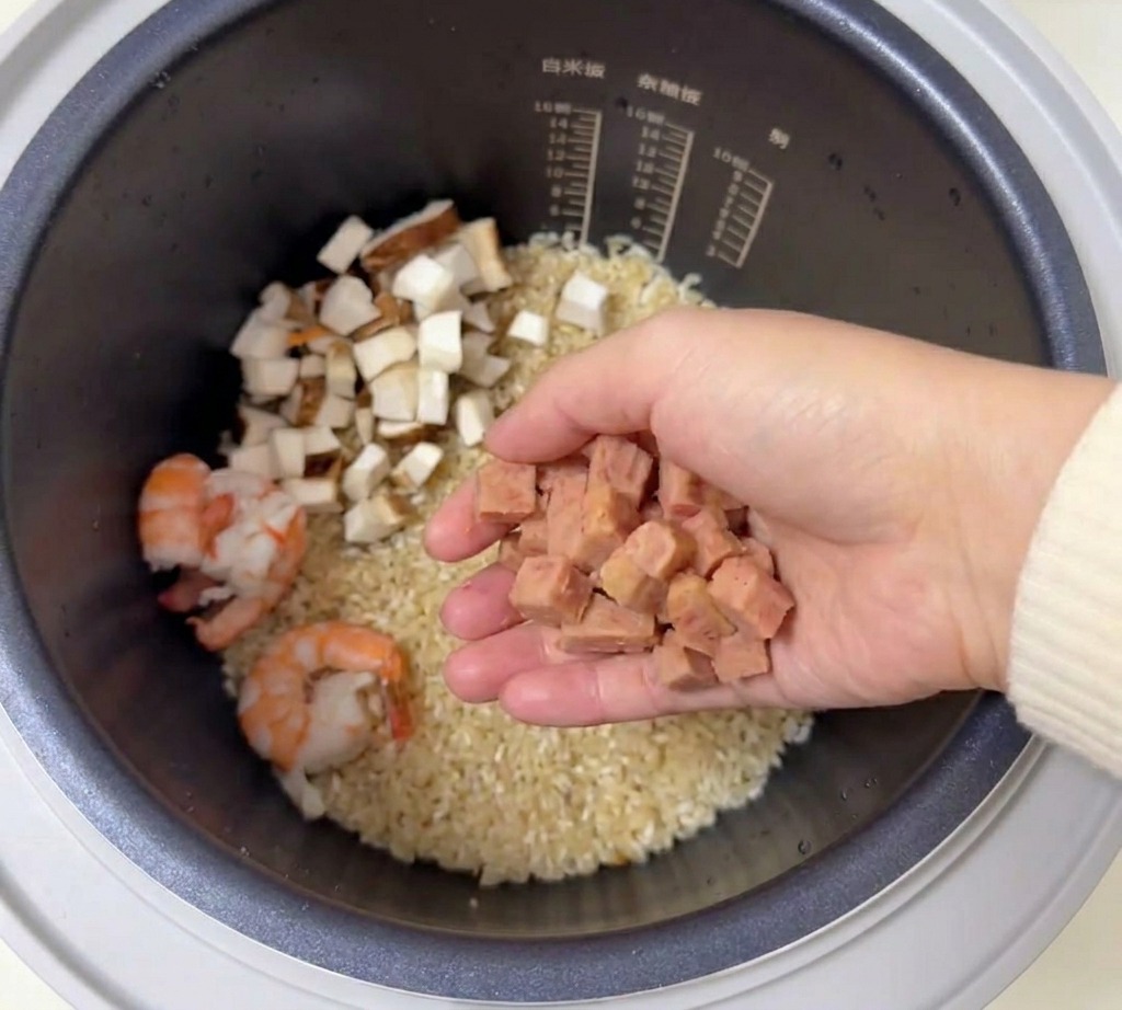 Una mano tiene una manciata di dadini di carne in scatola sopra una pentola per cuociriso con riso e gamberi.