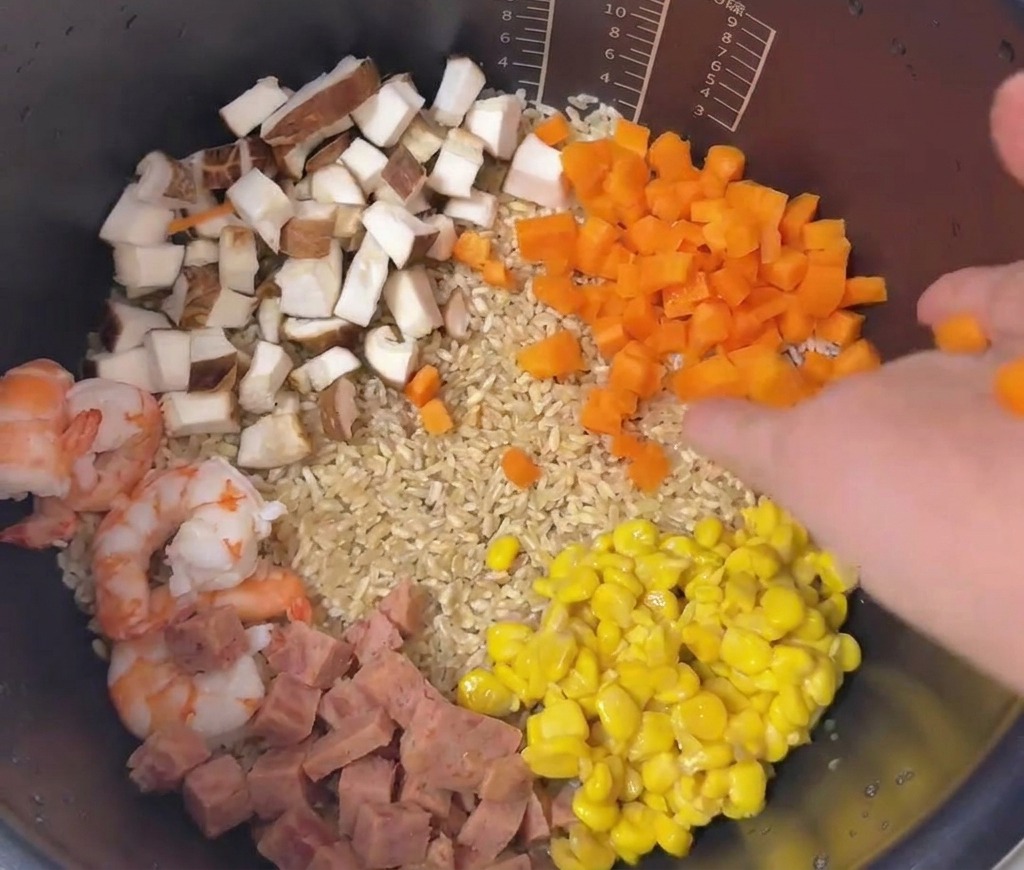 Una disposizione colorata di carote a dadini, mais, carne, funghi e gamberi stratificati sopra il riso in una pentola.