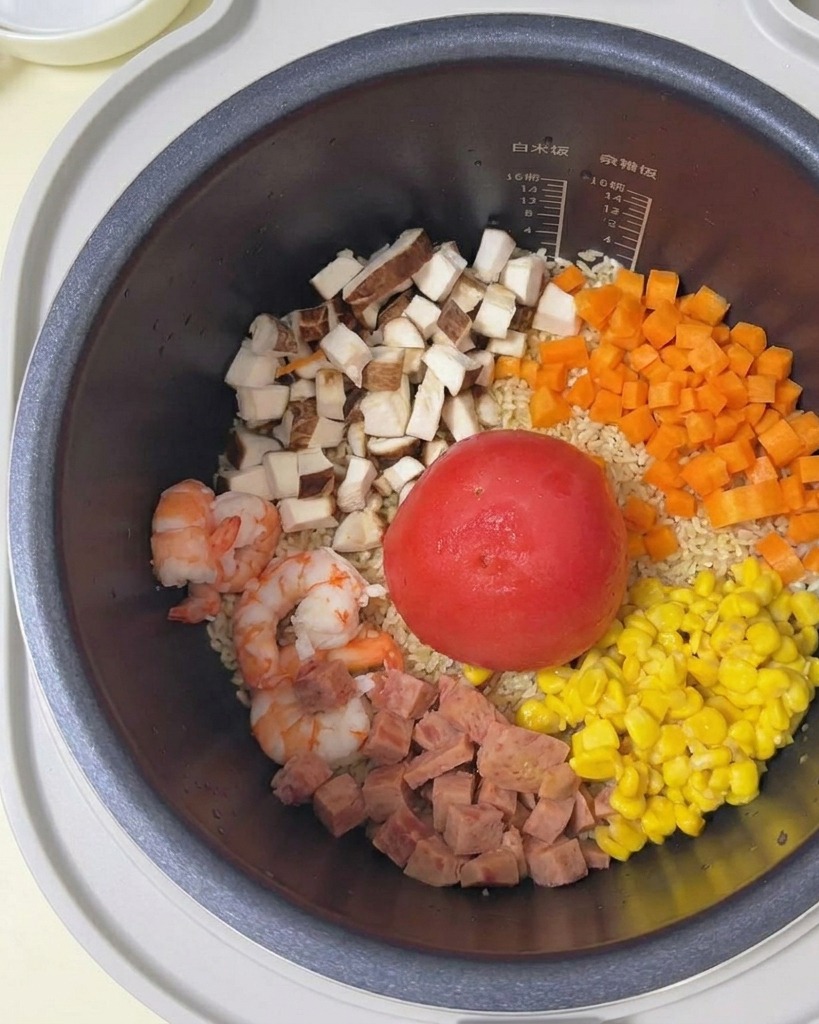 Acqua limpida versata da una ciotola bianca in un cuociriso pieno di riso, un pomodoro intero e vari ingredienti a dadini.