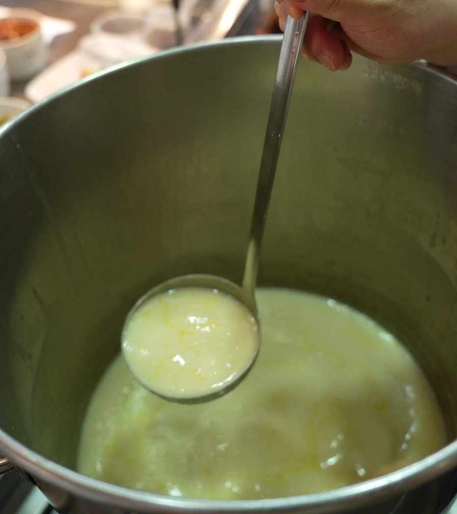 Un mestolo in acciaio che solleva un brodo di pollo denso, cremoso e dorato da una pentola professionale.