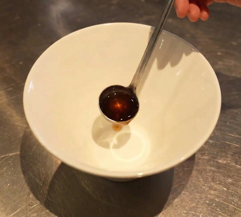 Un piccolo mestolo versa un condimento scuro sul fondo di una ciotola bianca vuota per ramen.