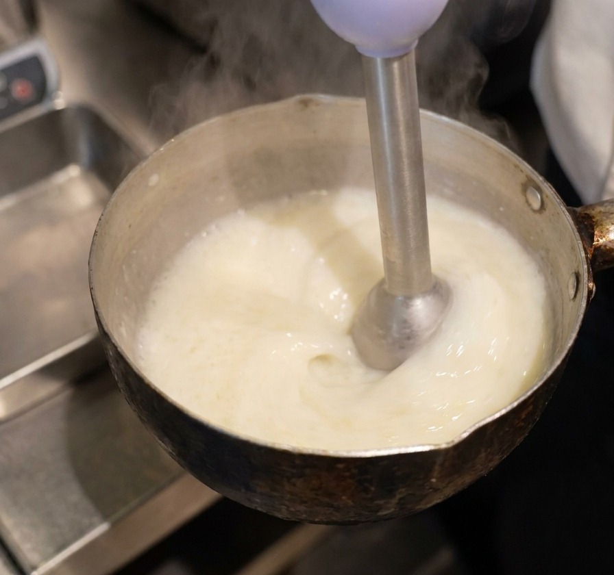 Un frullatore a immersione in un pentolino crea una schiuma densa e bianca sulla superficie del liquido giallo.