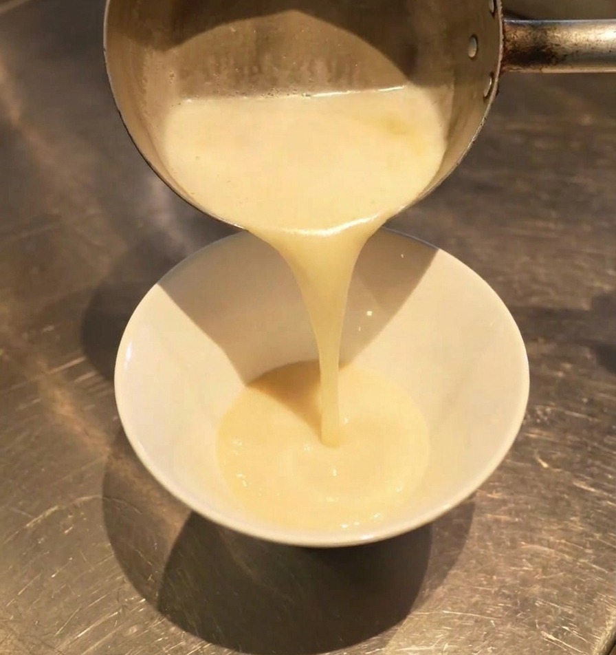 Brodo bianco e spumoso versato da un pentolino in una ciotola bianca, mescolandosi al condimento scuro sul fondo.