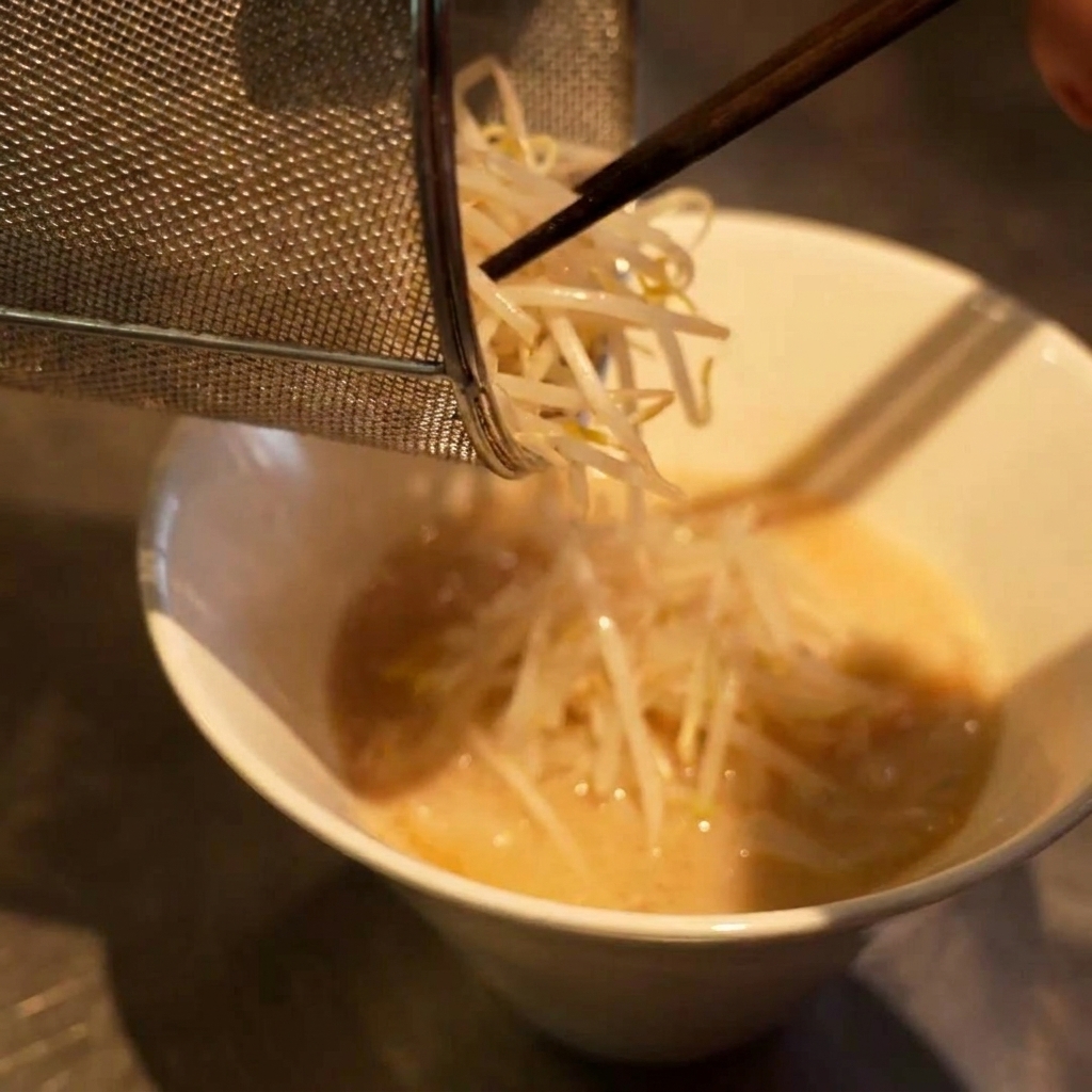 Un piccolo colino versa una manciata di germogli di soia bianchi sopra i noodles e il brodo.