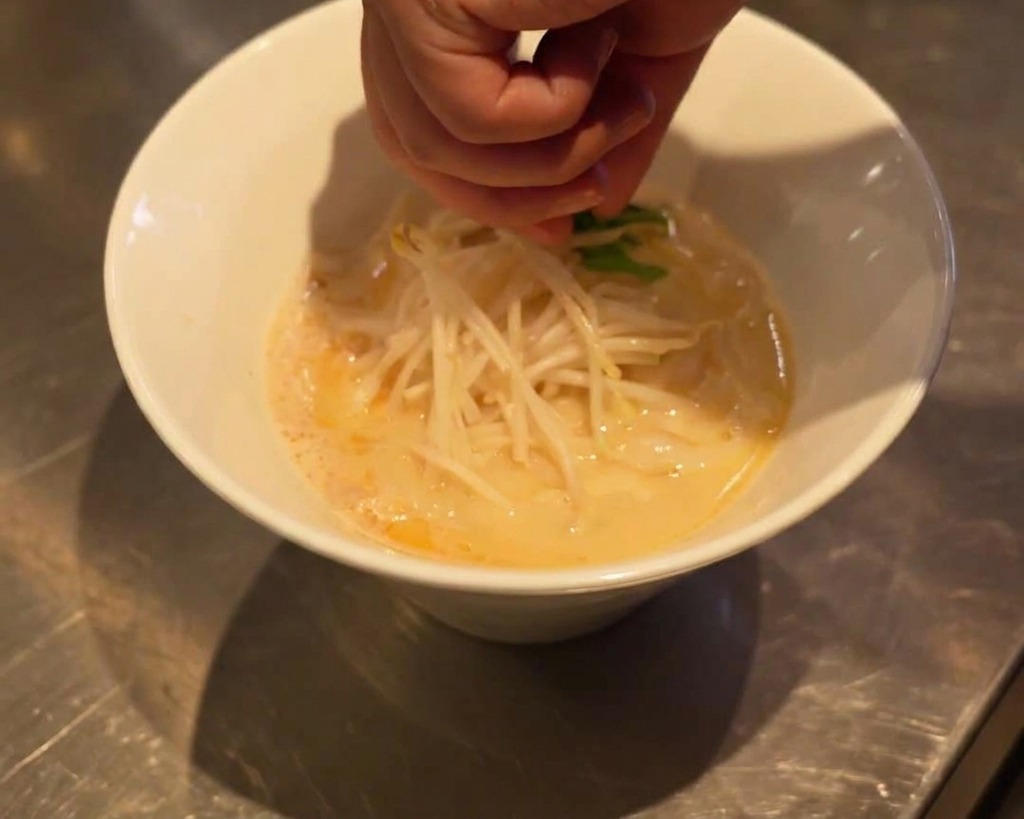 Una mano posiziona foglie di spinaci verdi sopra i germogli di soia in una ciotola di ramen.