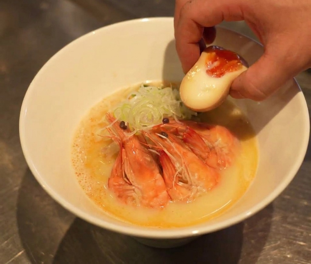 Una mano mette metà uovo sodo con il tuorlo cremoso in una ciotola di ramen quasi completa.