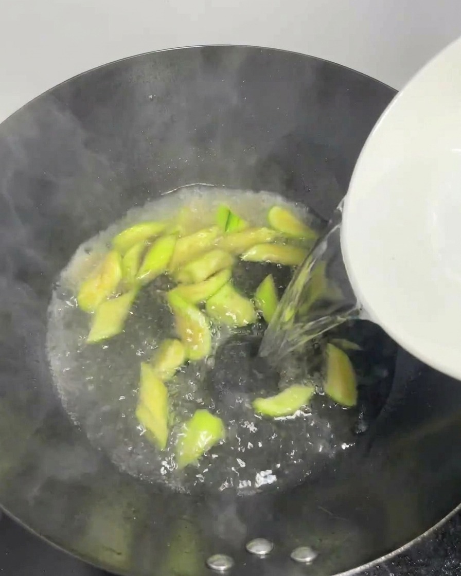 Acqua limpida versata da una ciotola bianca in un wok bollente con fette di luffa.
