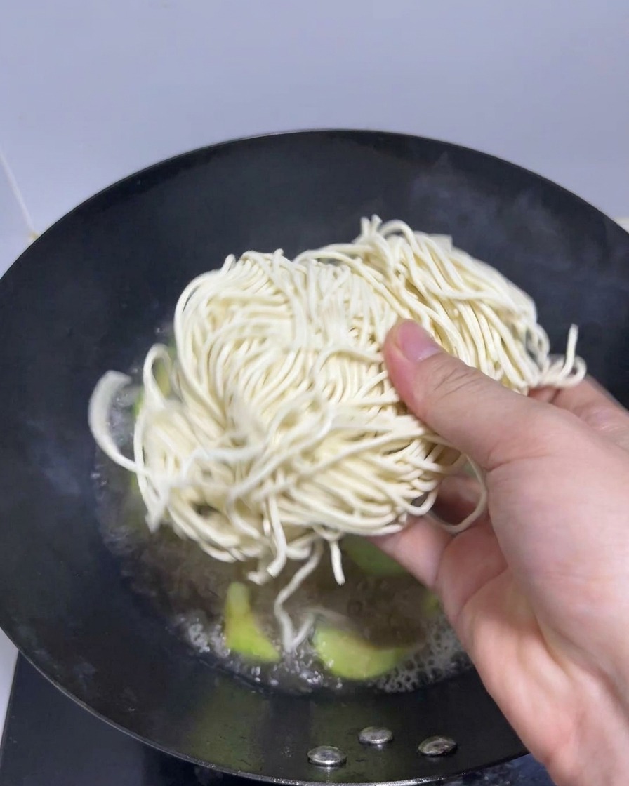Una mano che mette un nido di noodles freschi per ramen in un wok con brodo bollente.