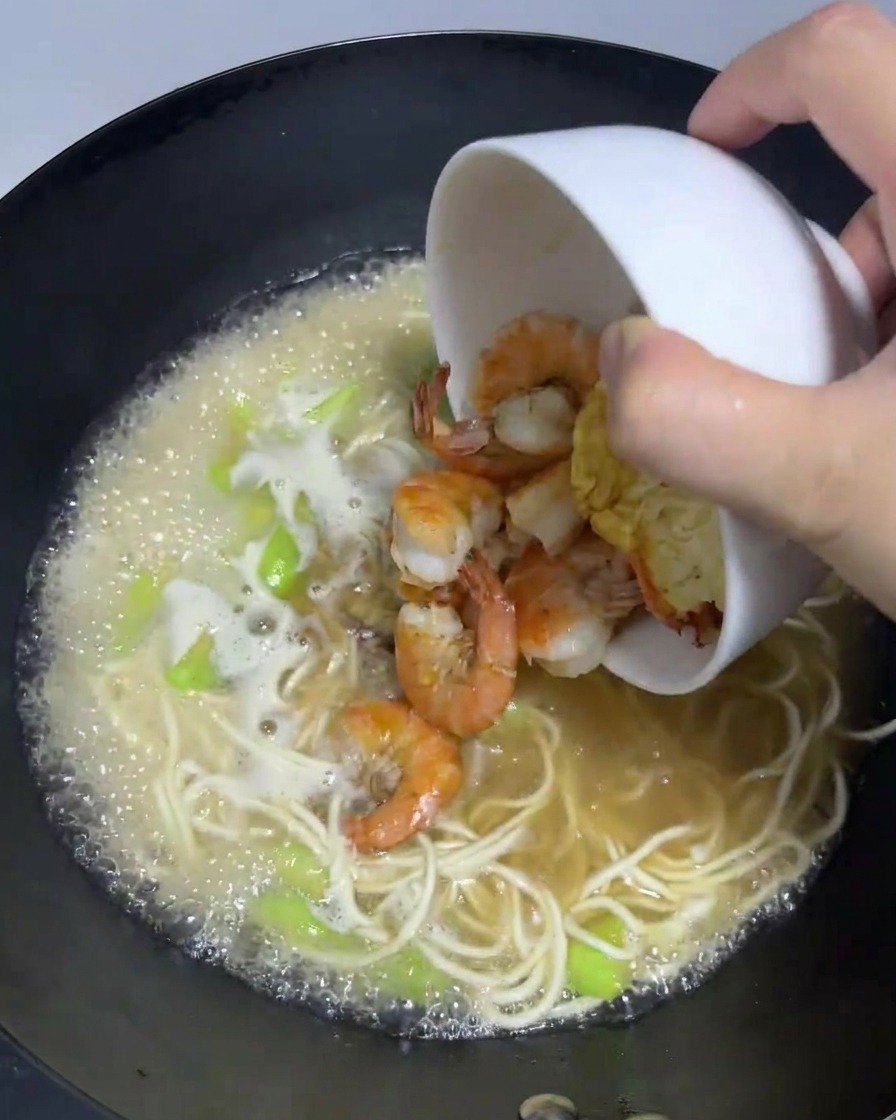 Gamberi cotti e un uovo fritto versati da una ciotola in un wok con ramen che sobbolle.