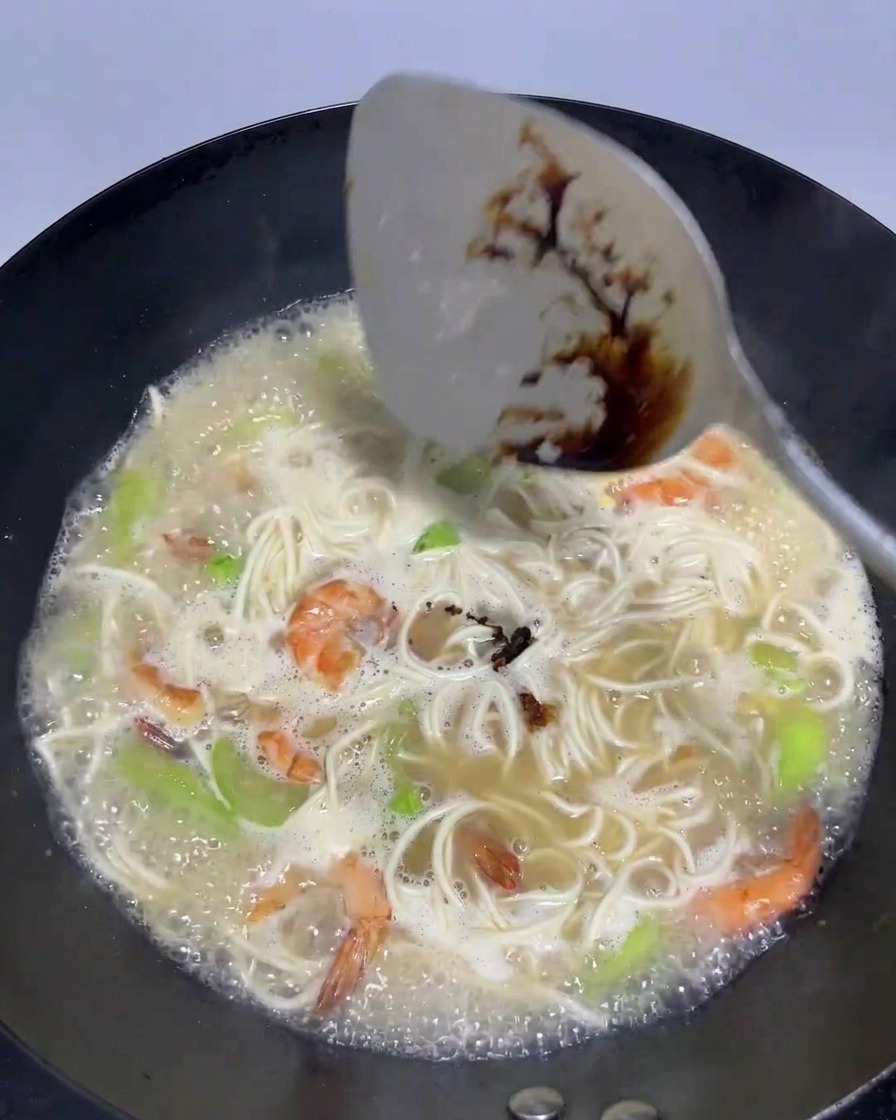 Una spatola che versa un filo di salsa di soia scura in un wok con brodo bollente, noodles, gamberi e luffa.