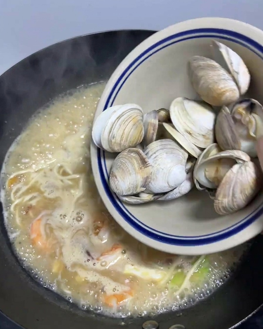 Una ciotola di vongole sbollentate versata in un grande wok pieno di ramen ai frutti di mare fumante.