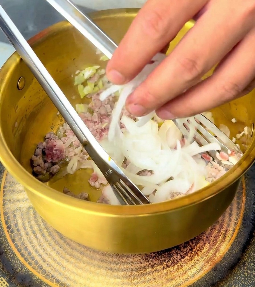 Una mano aggiunge cipolle bianche affettate in una pentola dorata con carne e aromi.