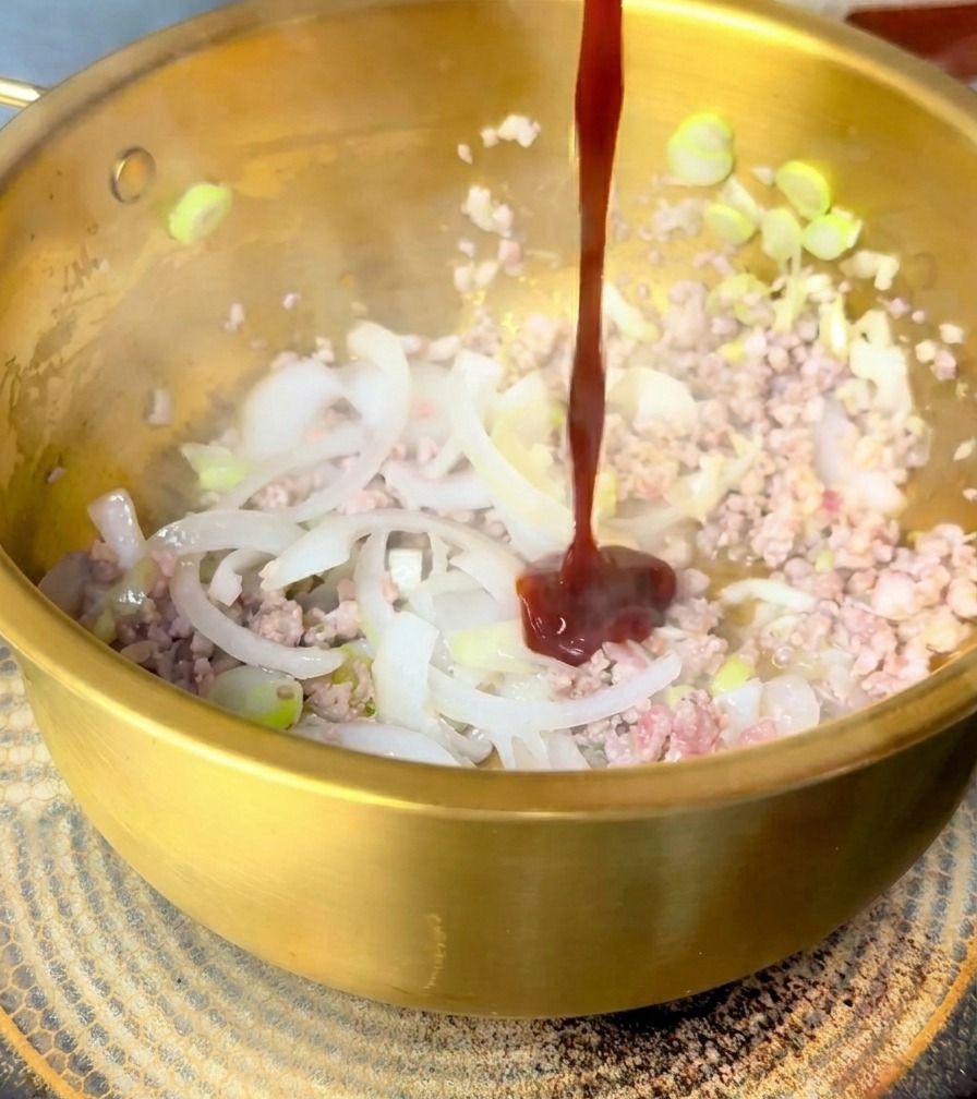Un getto denso di salsa rossa versato sul maiale e le cipolle in una pentola dorata.