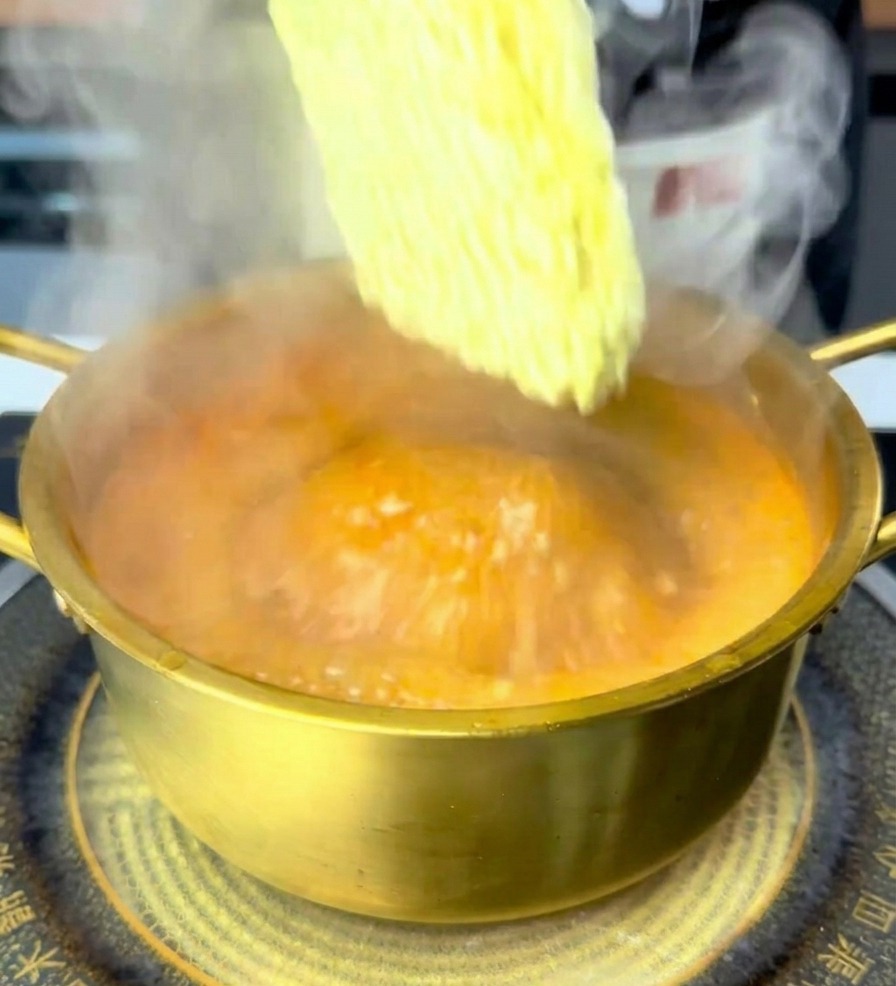 Una mano inserisce un panetto di noodle istantanei in una pentola con brodo rosso bollente e fumante.