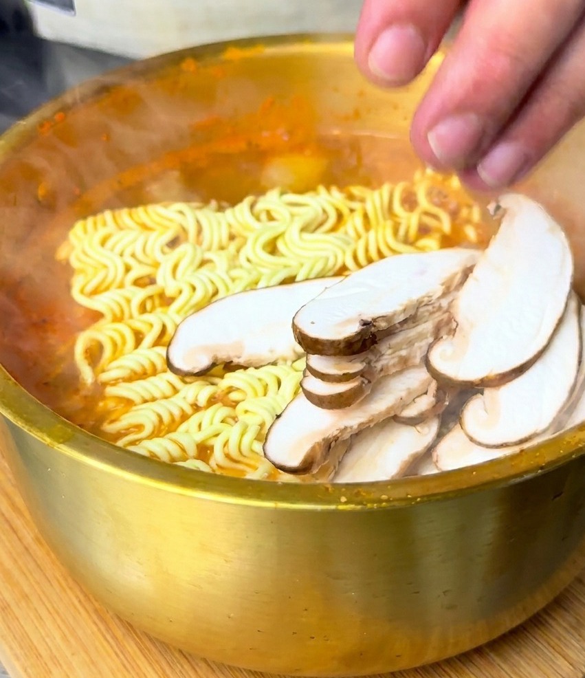 Una mano dispone funghi shiitake affettati sopra i noodle immersi nel brodo rosso.