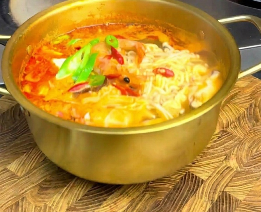 Ramen ai frutti di mare piccante in pentola dorata con fettine di peperoncino su un tagliere di legno.