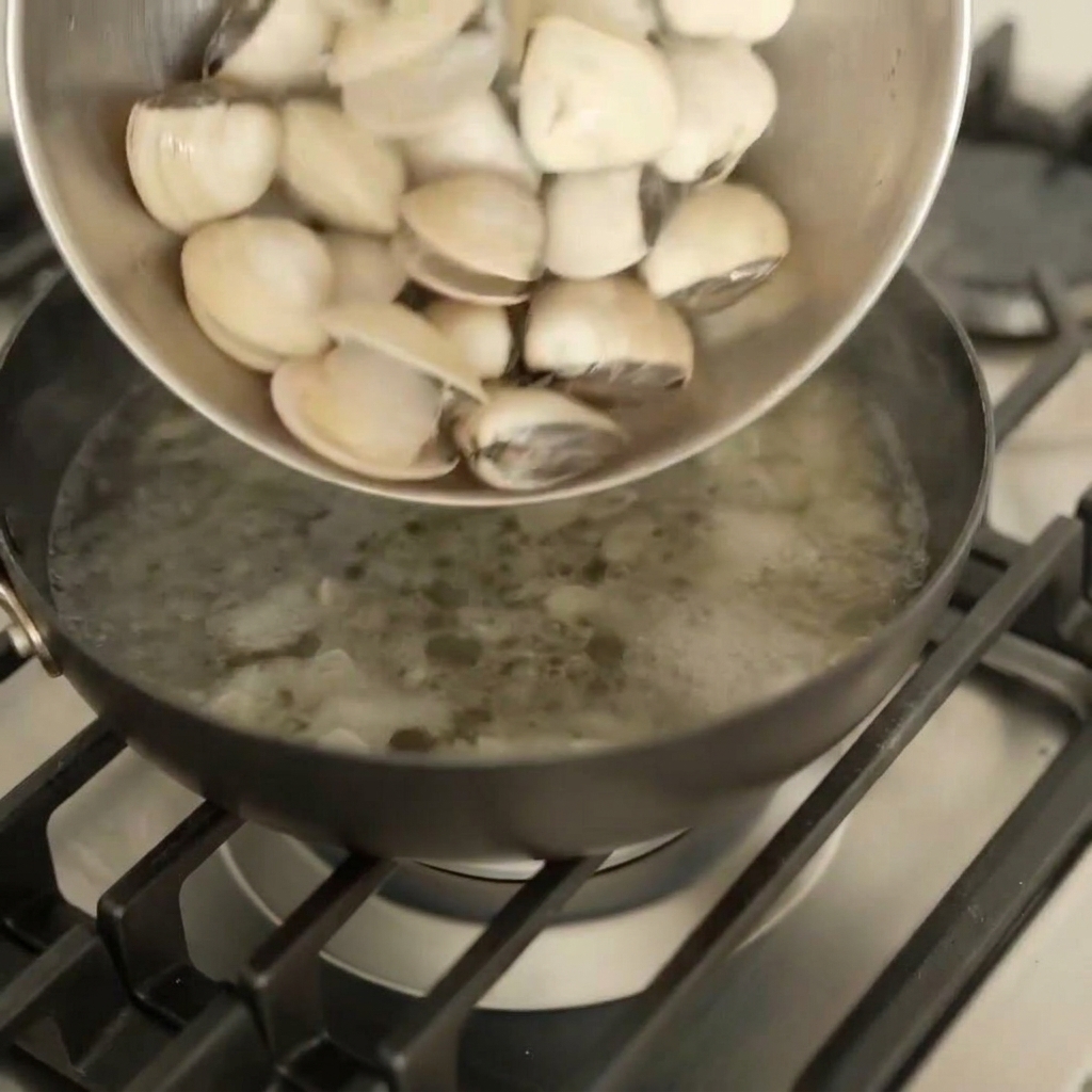Vongole fresche versate da una ciotola di metallo in una padella scura sul fuoco.