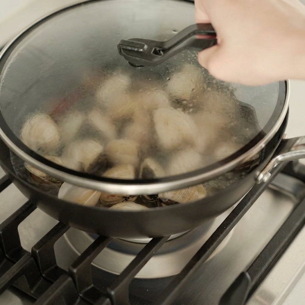 Una mano posiziona un coperchio di vetro sopra la padella con le vongole per cuocerle a vapore.