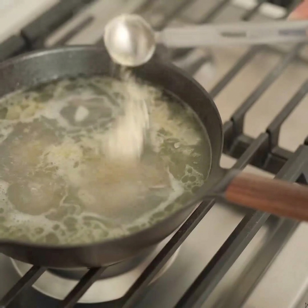 Un cucchiaio dosatore dargento versa un liquido scuro in una pentola di brodo chiaro per ramen.