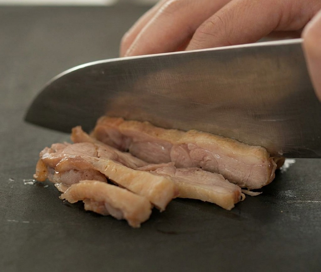 Un coltello affilato affetta un pezzo di anatra rosolata e cotta su un tagliere scuro.