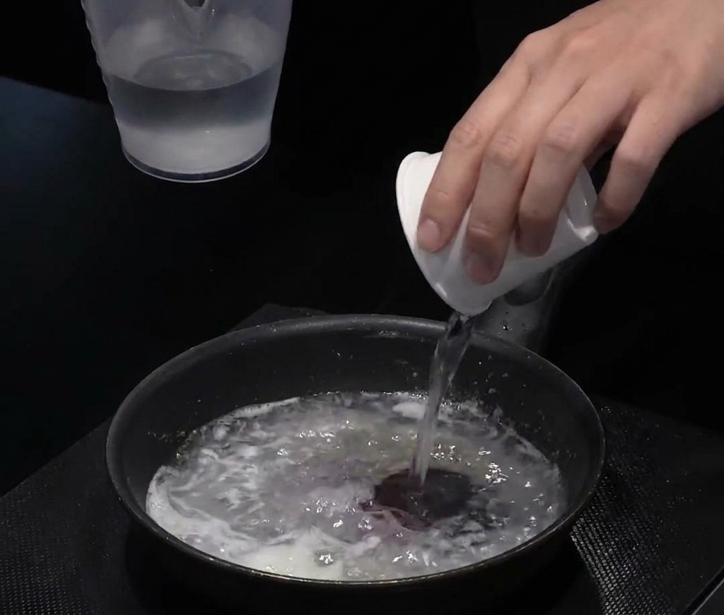 Acqua versata da una tazzina bianca in una padella nera contenente il mix di olio e farina.
