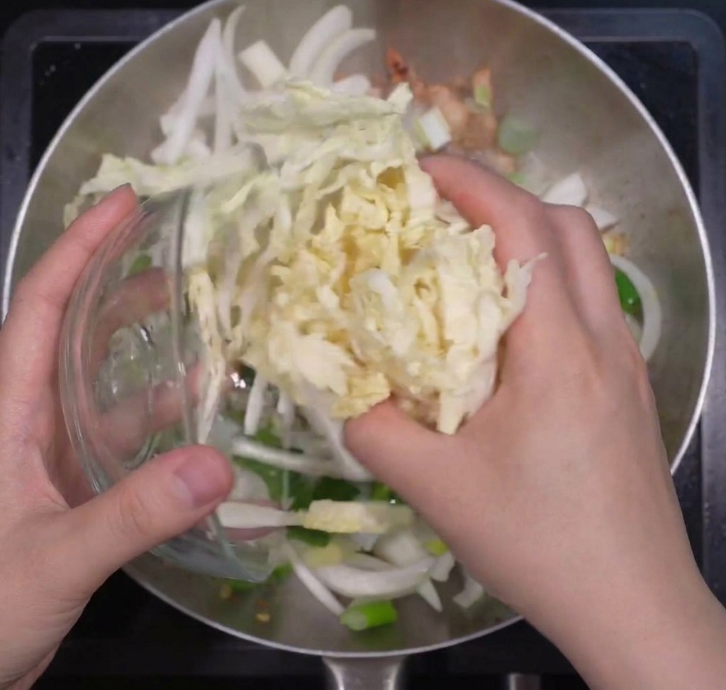 Mani che aggiungono cavolo cinese sminuzzato in una padella con cipolle e cipollotti già rosolati.