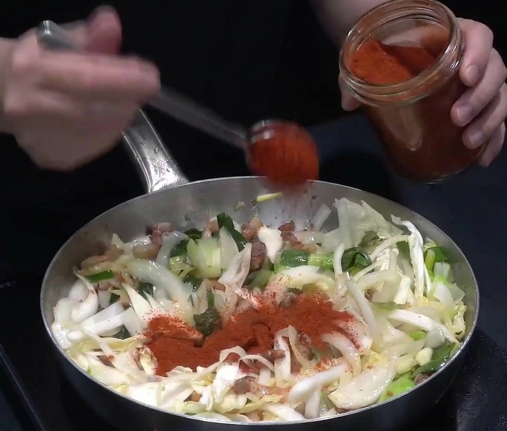 Peperoncino rosso in polvere versato con un cucchiaio su verdure e pancetta saltate in una padella capiente.