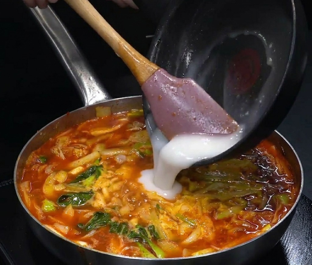 Un liquido bianco opaco versato da un pentolino nero in una pentola di zuppa rossa bollente.