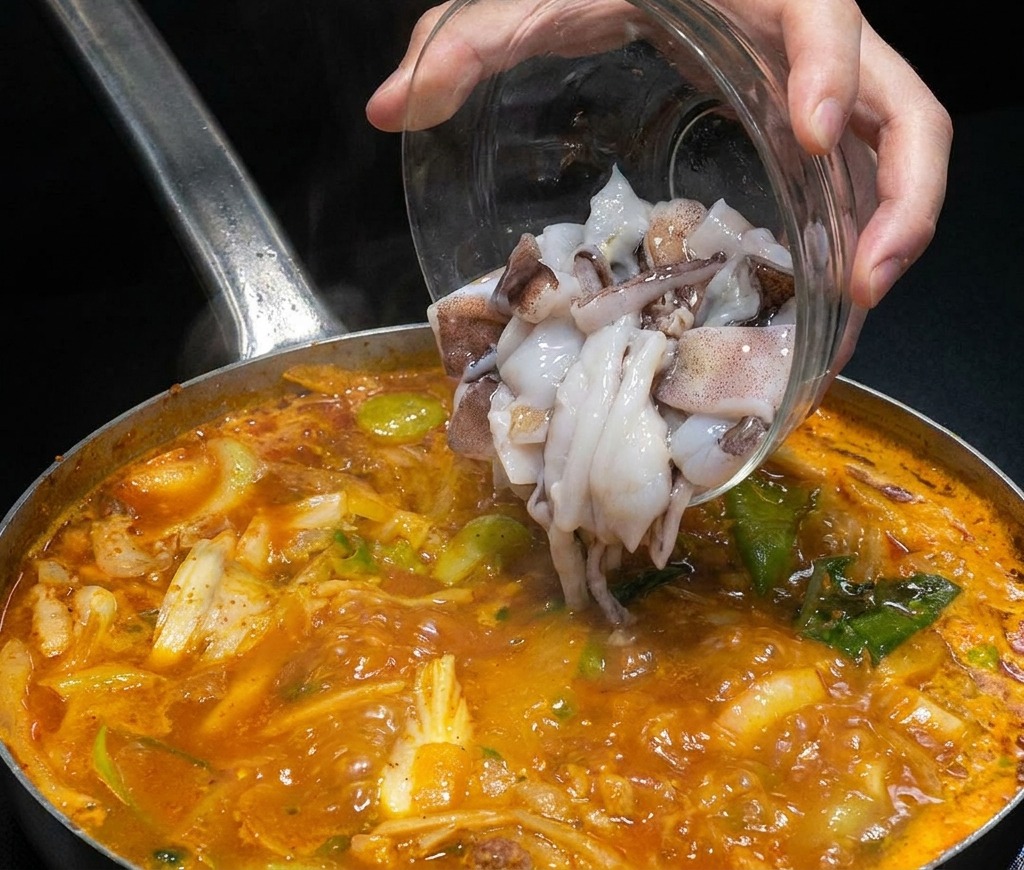 Pezzi di calamaro crudo versati da una ciotola di vetro in una pentola fumante di brodo rosso di mare.