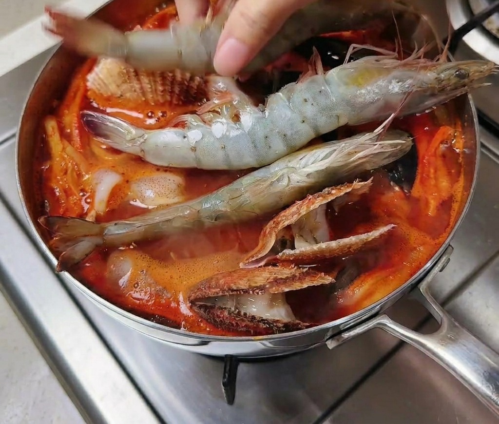 Mano che aggiunge gamberoni crudi in una pentola di zuppa rossa piccante con molluschi.