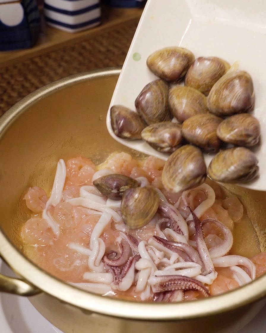 Vongole fresche versate da un piatto bianco in una pentola dorata contenente gamberi e anelli di calamaro saltati.