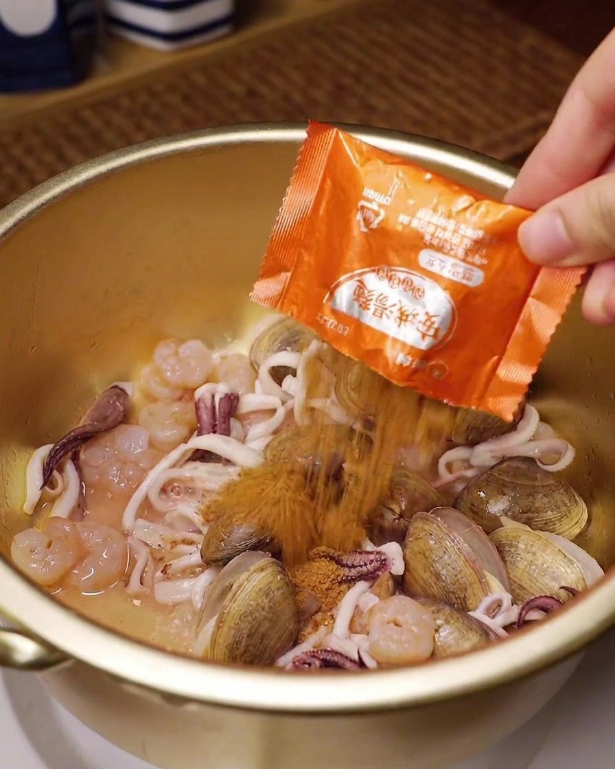 Una mano versa il condimento del ramen istantaneo su vongole, calamari e gamberi in una pentola di ottone.