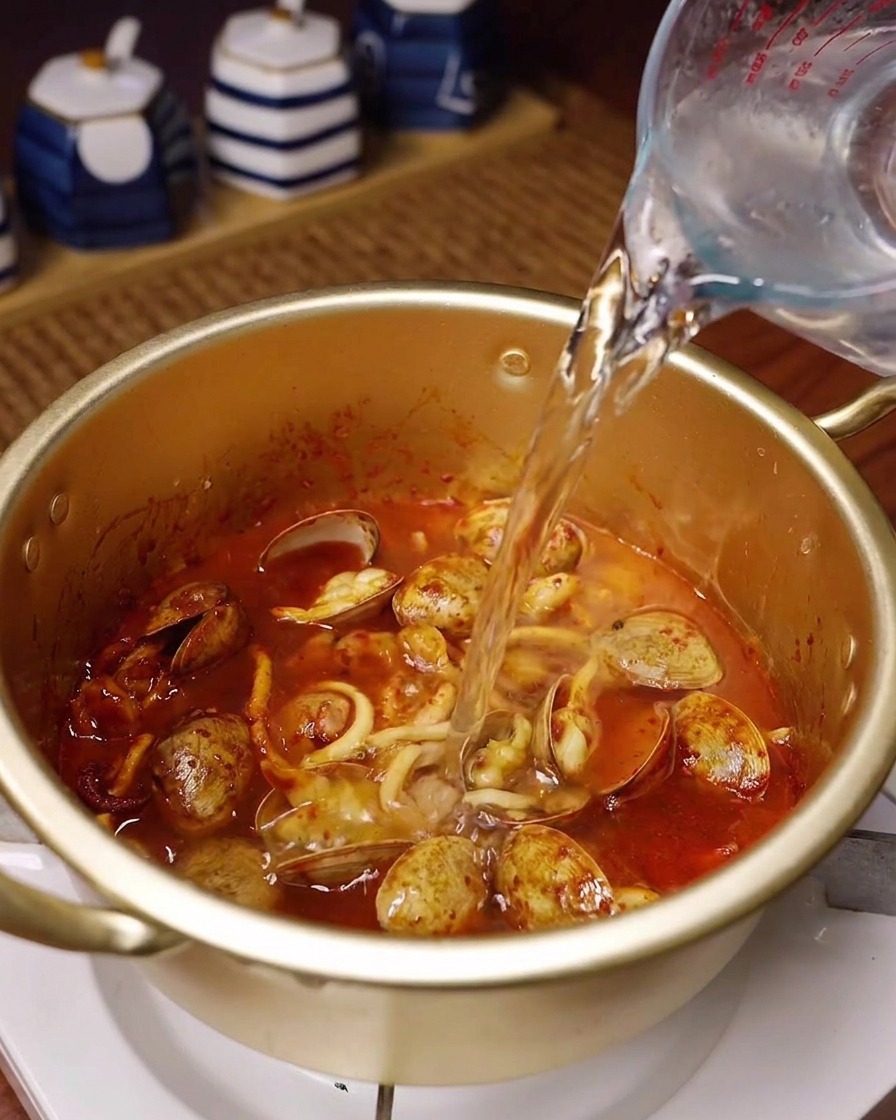 Acqua fresca versata da una caraffa graduata in una pentola dorata con i frutti di mare speziati.