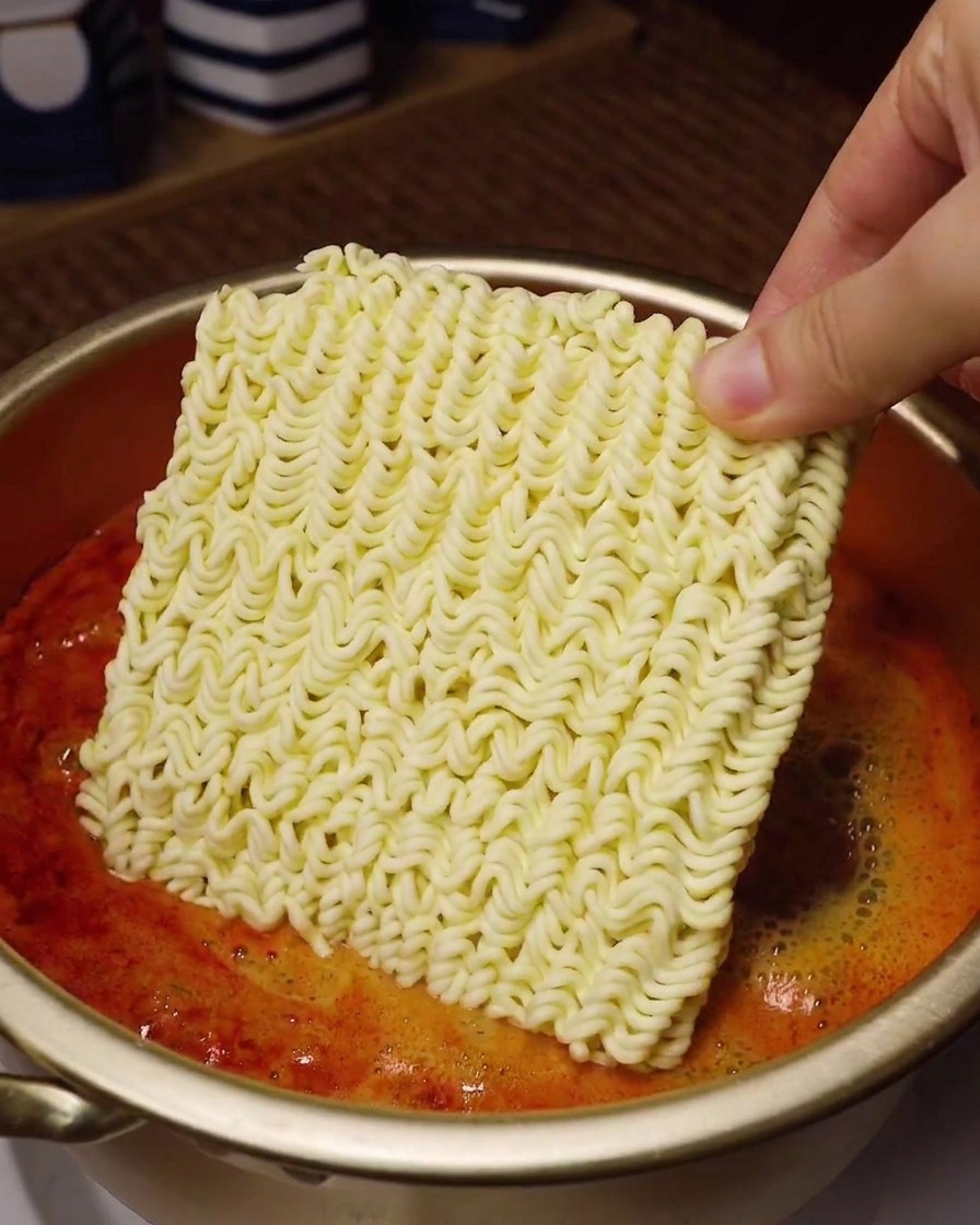 Una mano tiene il blocco di noodles istantanei sopra una pentola con brodo rosso bollente.