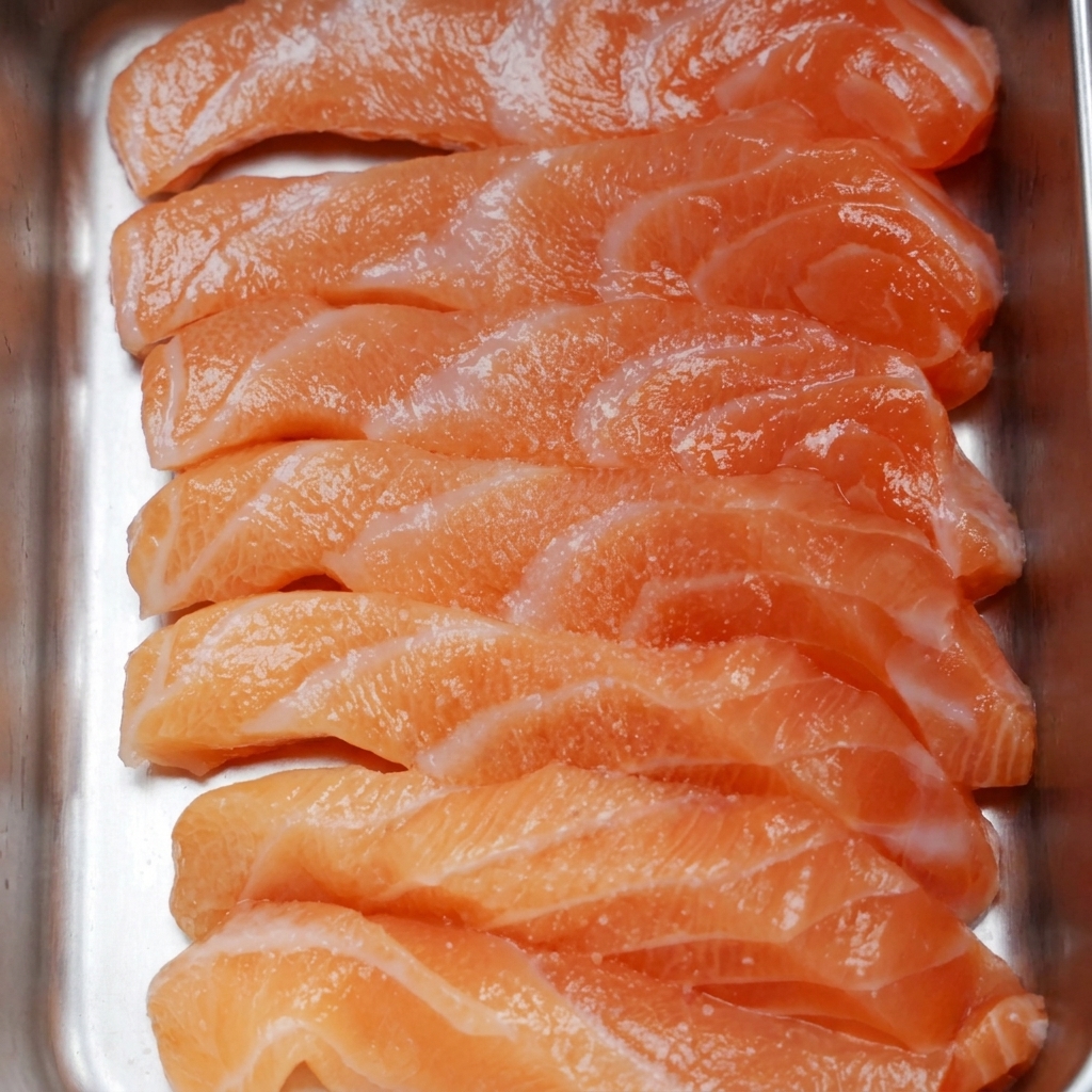 Fette di salmone fresco crudo disposte in un vassoio di metallo, cosparse di sale fino e granelli di pepe nero.
