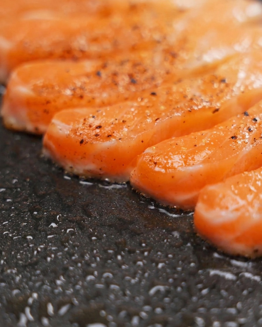 Filetti di salmone conditi che sfrigolano in una padella antiaderente scura, mostrando una doratura visibile.